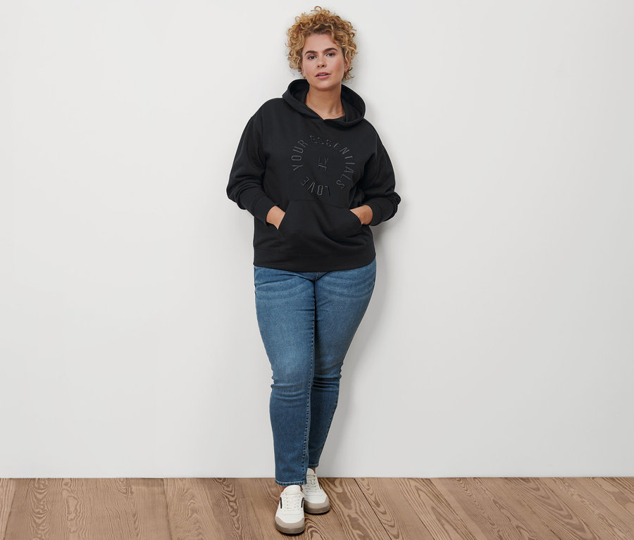 Une femme pose, portant un sweat-shirt à capuche noir, un jean slim en denim « Fit Emma » bleu moyen et des baskets en cuir.