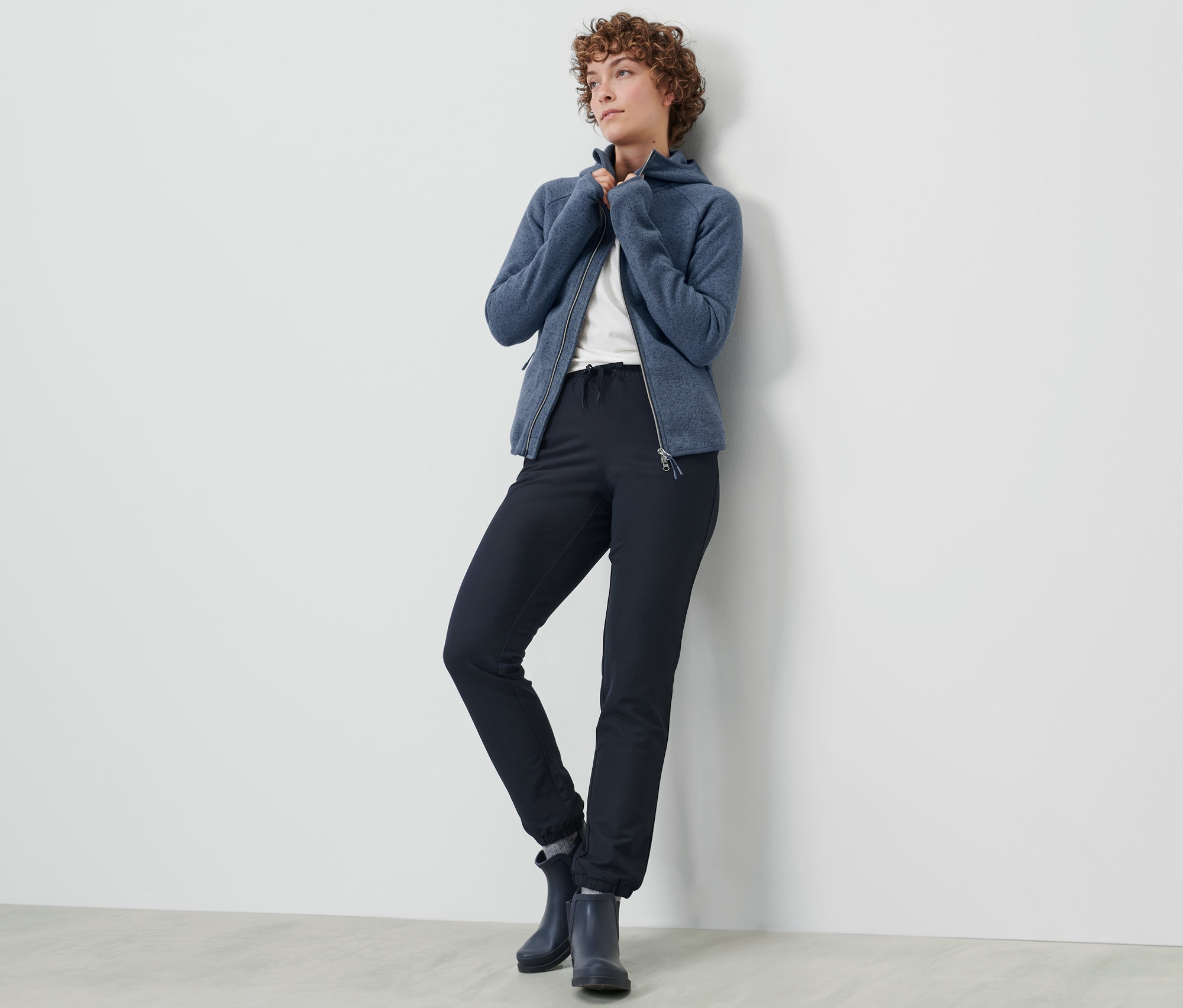 Une femme pose en pantalon de jogging en softshell et veste zippée bleue contre un mur.