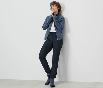 Une femme pose en pantalon de jogging en softshell et veste zippée bleue contre un mur.