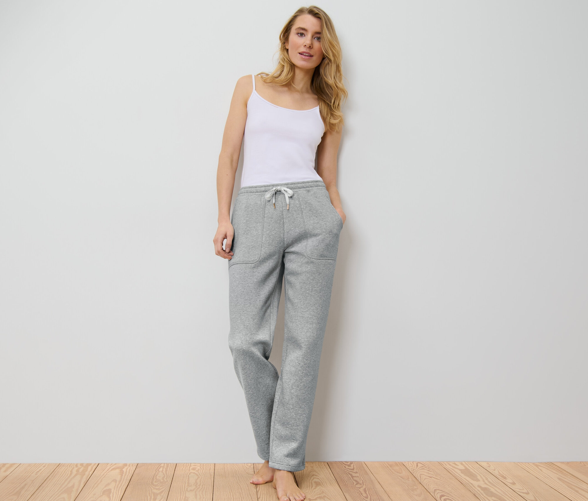 Femme en débardeur blanc et pantalon d’intérieur gris debout contre un mur blanc.