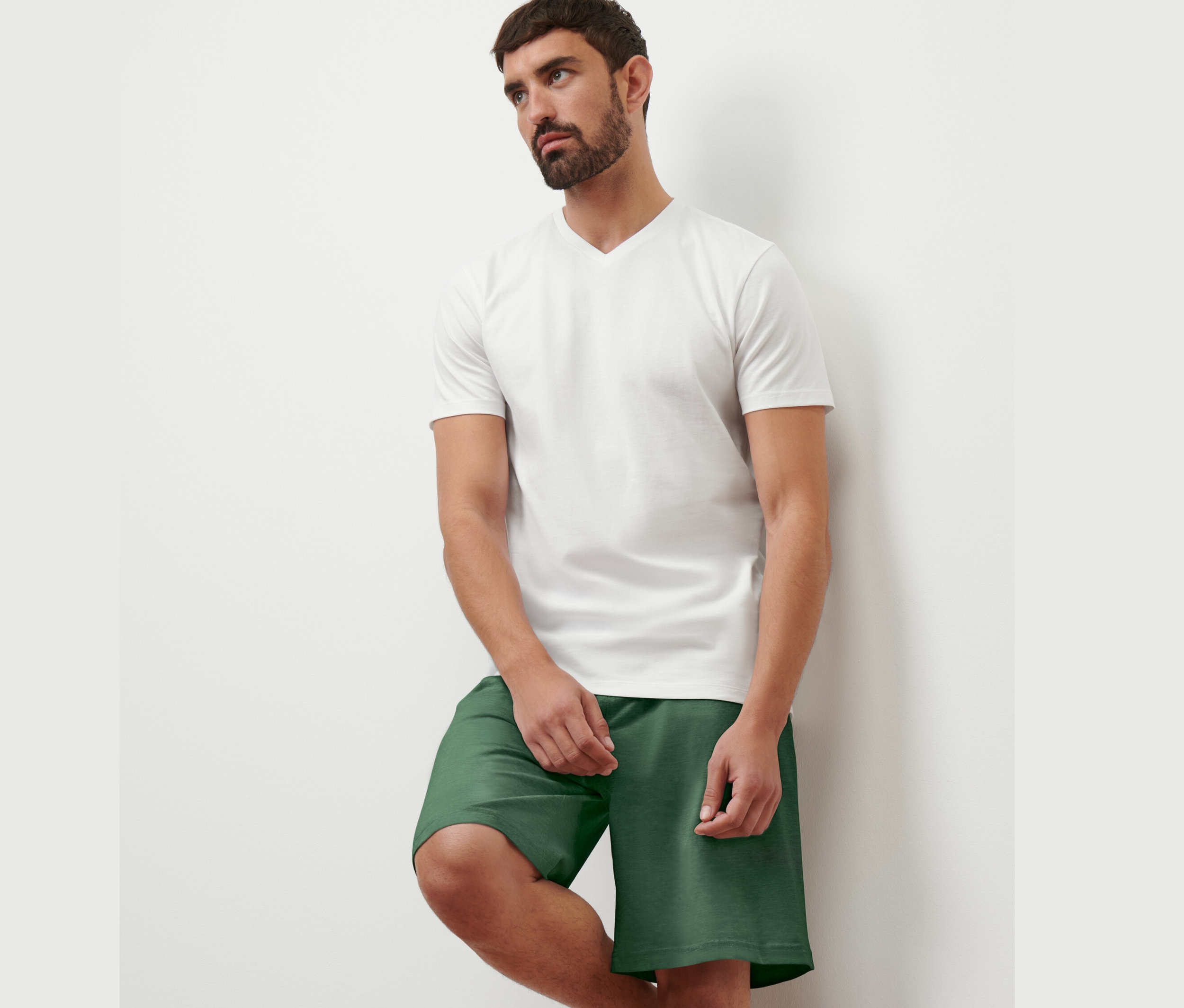 Un homme appuyé contre un mur porte un t-shirt blanc à col en V et un short vert. Il porte 1 des 2 t-shirts à col en V.