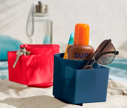 Deux supports à gobelets de plage dans le sable. Dans le support bleu se trouvent des lunettes de soleil et de la crème solaire.