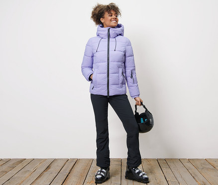 Femme en veste de ski matelassée, pantalon noir et chaussures de ski tenant un casque.