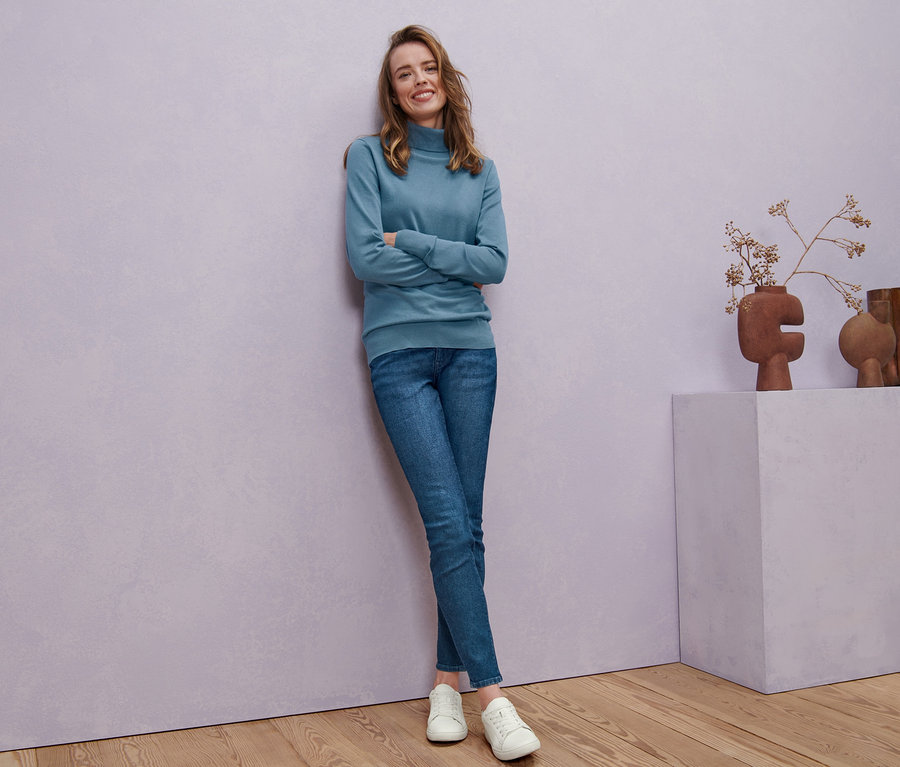 Femme posant en pull à col roulé en maille fine bleu fumée, jean denim skinny « Fit Hanna » et baskets en cuir.