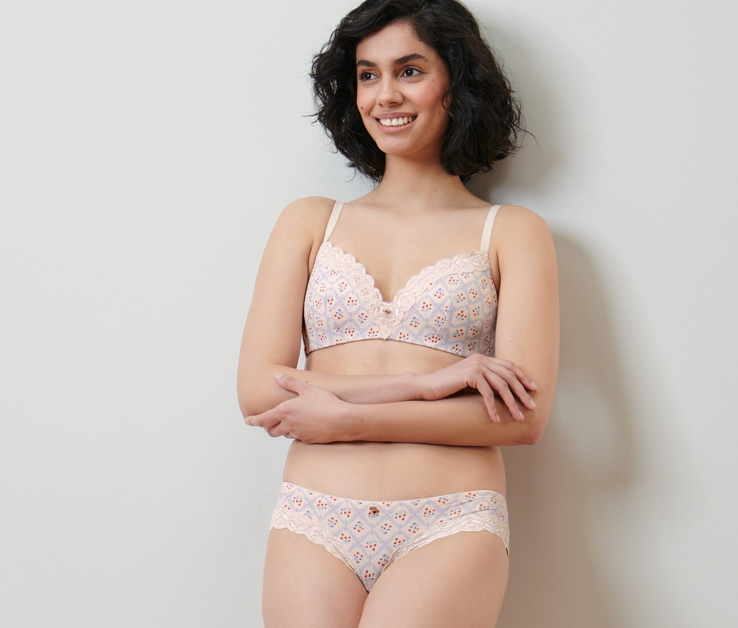 Une femme souriante pose dans un ensemble de sous-vêtements crème avec imprimé intégral et dentelle. Elle porte 2 slips brésiliens.