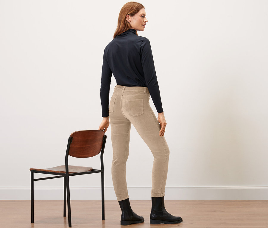 Une femme se tient de côté et s'appuie sur une chaise, elle porte un pantalon en velours camel.