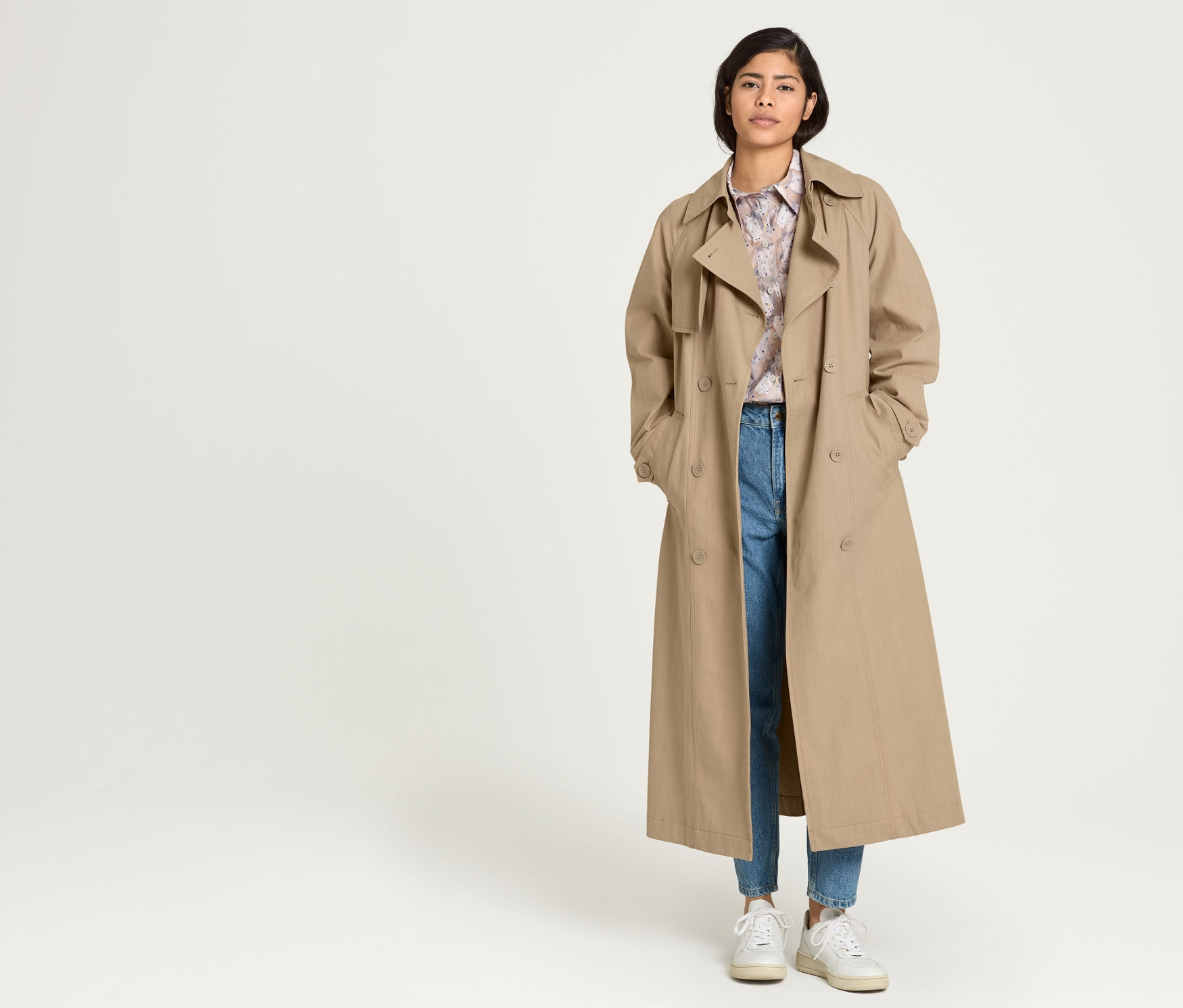 Une femme se tient debout, les mains dans les poches d'un trench-coat beige NAH/STUDIO en coton recyclé, sous lequel elle porte une blouse longue imprimée NAH/STUDIO en coton bio et un jean Mom bleu moyen NAH/STUDIO en coton recyclé.
