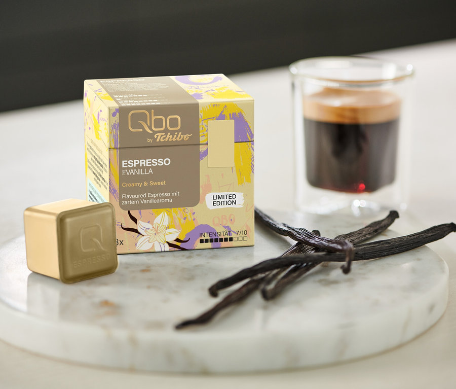 Une boîte de Qbo Flavoured Espresso Vanilla – 8 capsules, une capsule, un expresso et des gousses de vanille sur un plateau en marbre à côté d'un verre.