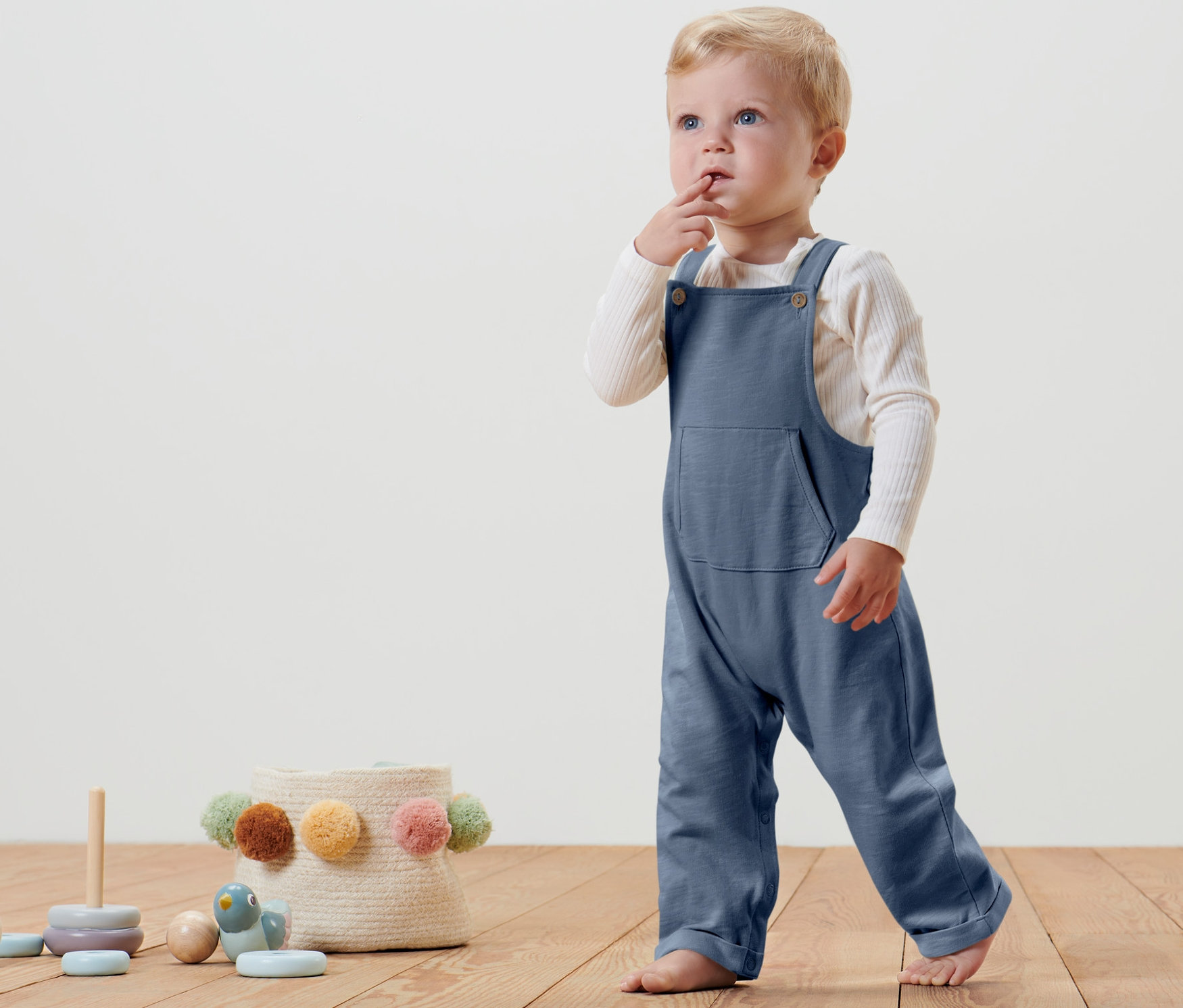 Un petit enfant se tient sur un plancher en bois et porte une salopette bleue pour bébé.