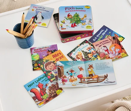 Sur une table blanche sont posés une boîte à livres ouverte « La boîte de Noël colorée de Pixi » et plusieurs livres pour enfants, à côté d'un gobelet bleu contenant des crayons de couleur.