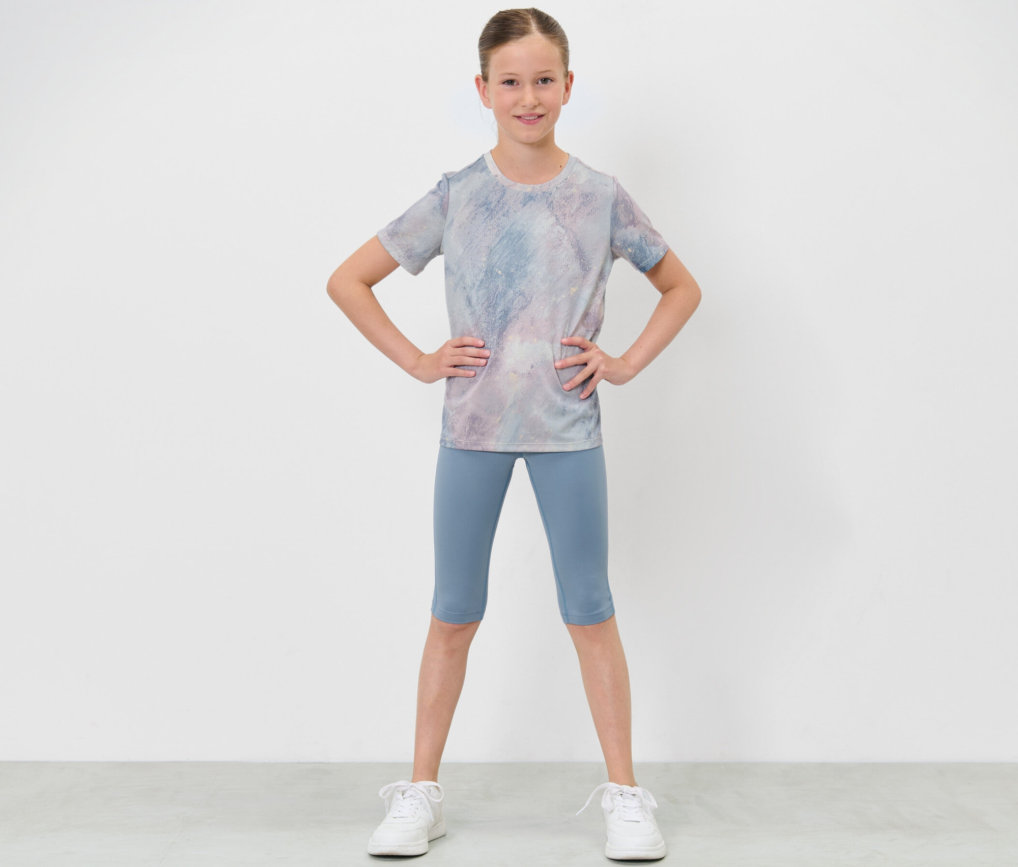 Une fille en tenue de sport pose les mains sur les hanches, portant un corsaire de sport pour enfants et un t-shirt.