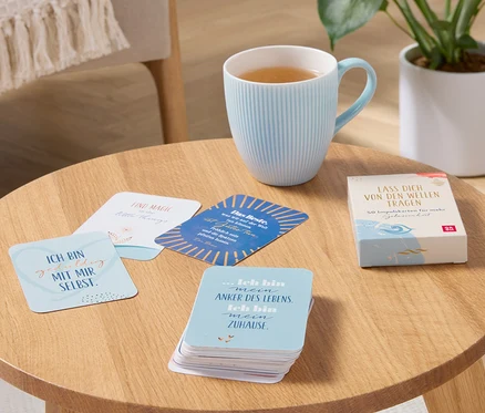 Sur une table en bois se trouvent une tasse bleu clair avec une boisson et un jeu de cartes de sérénité « Laisse-toi porter par les vagues ».