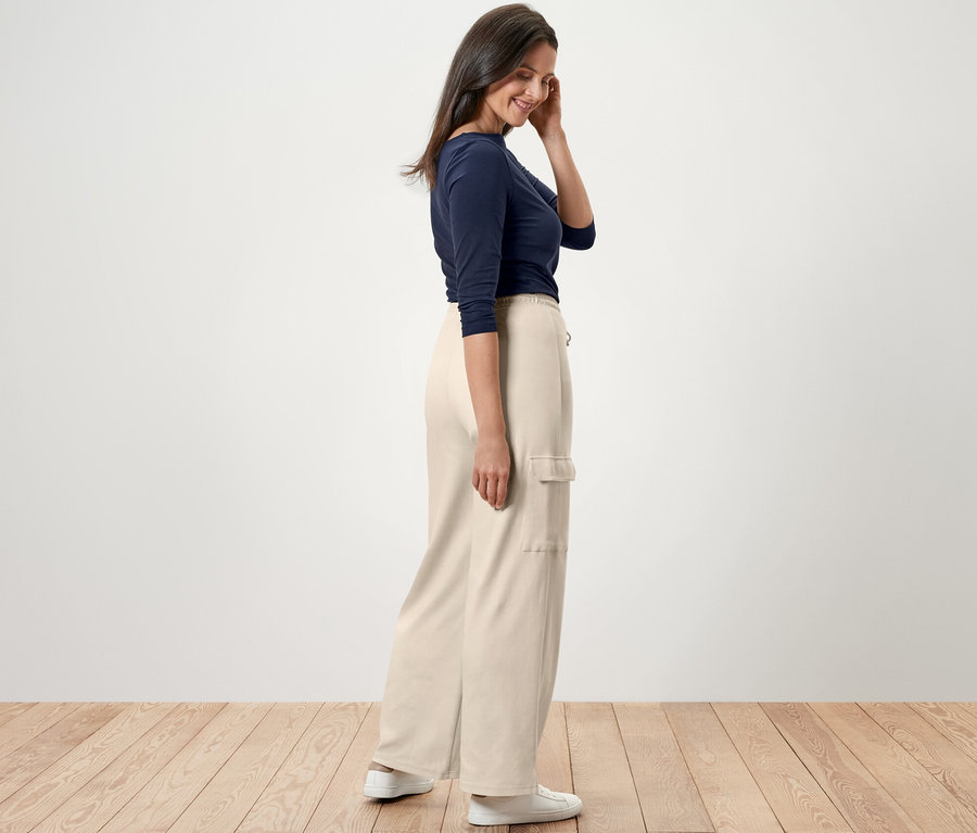 Une femme debout de profil porte un pantalon molletonné blanc crème avec des poches cargo, un t-shirt bleu et des baskets blanches.