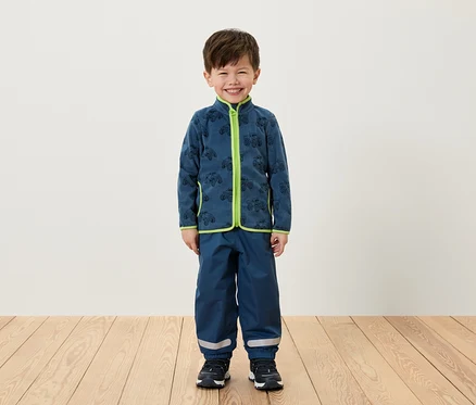 Un garçon debout portant une veste en polaire bleue avec des bordures vertes et un pantalon bleu.