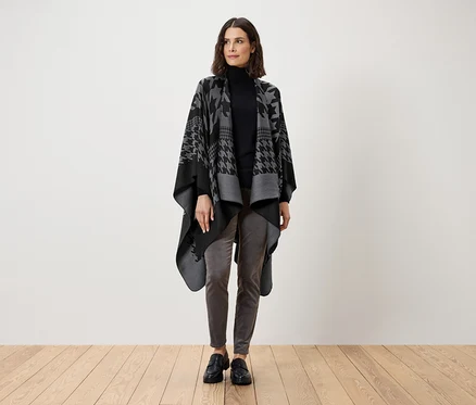 Une femme debout portant un col roulé noir, un pantalon gris et une cape pied-de-poule grise et noire.