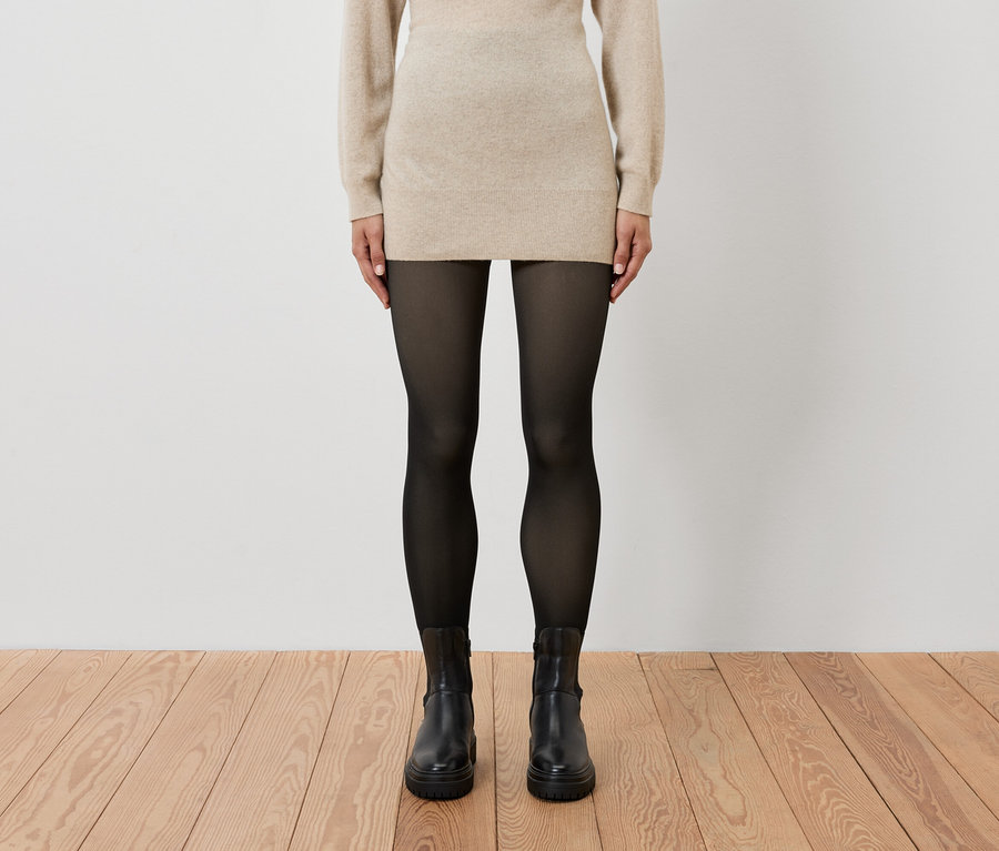 Une femme porte une robe beige, des collants noirs et des bottes noires.