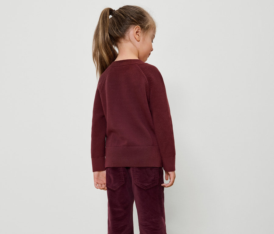 Fillette avec une queue de cheval portant un pull bordeaux et un pantalon en velours, vue de dos.