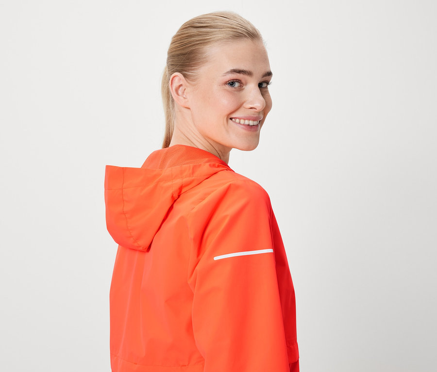 Une femme souriant par-dessus son épaule porte une veste orange à capuche avec une bande réfléchissante.
