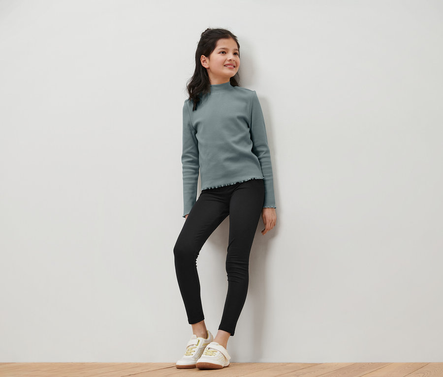 Une jeune fille souriante portant un haut à manches longues bleu-vert, un jean noir et des baskets blanches pose devant un mur blanc.