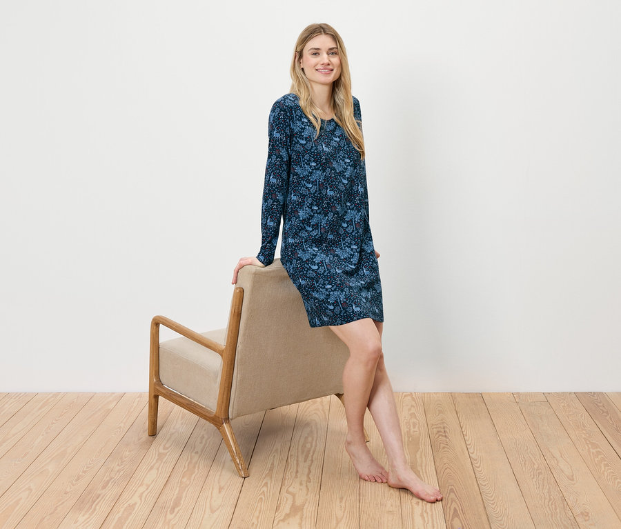 Une femme est assise, appuyée contre un fauteuil beige, et porte une robe fleurie bleue.