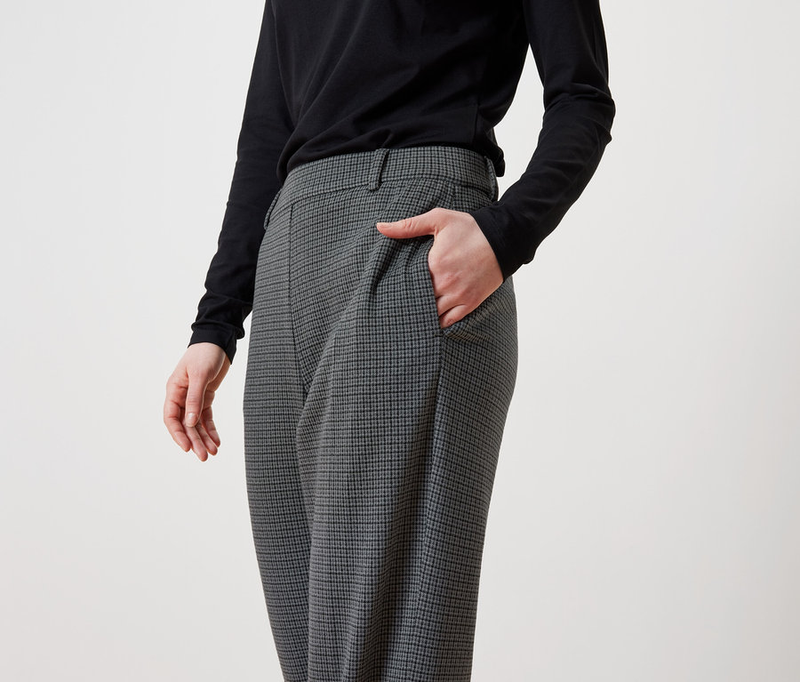 Une femme a sa main dans la poche d'un pantalon gris à carreaux et porte un t-shirt noir à manches longues.