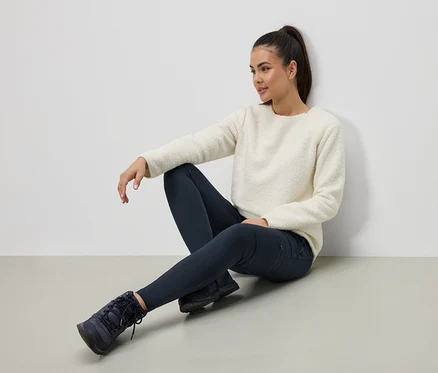 Une femme est assise par terre et porte un pull crème, un legging bleu marine et des bottes.