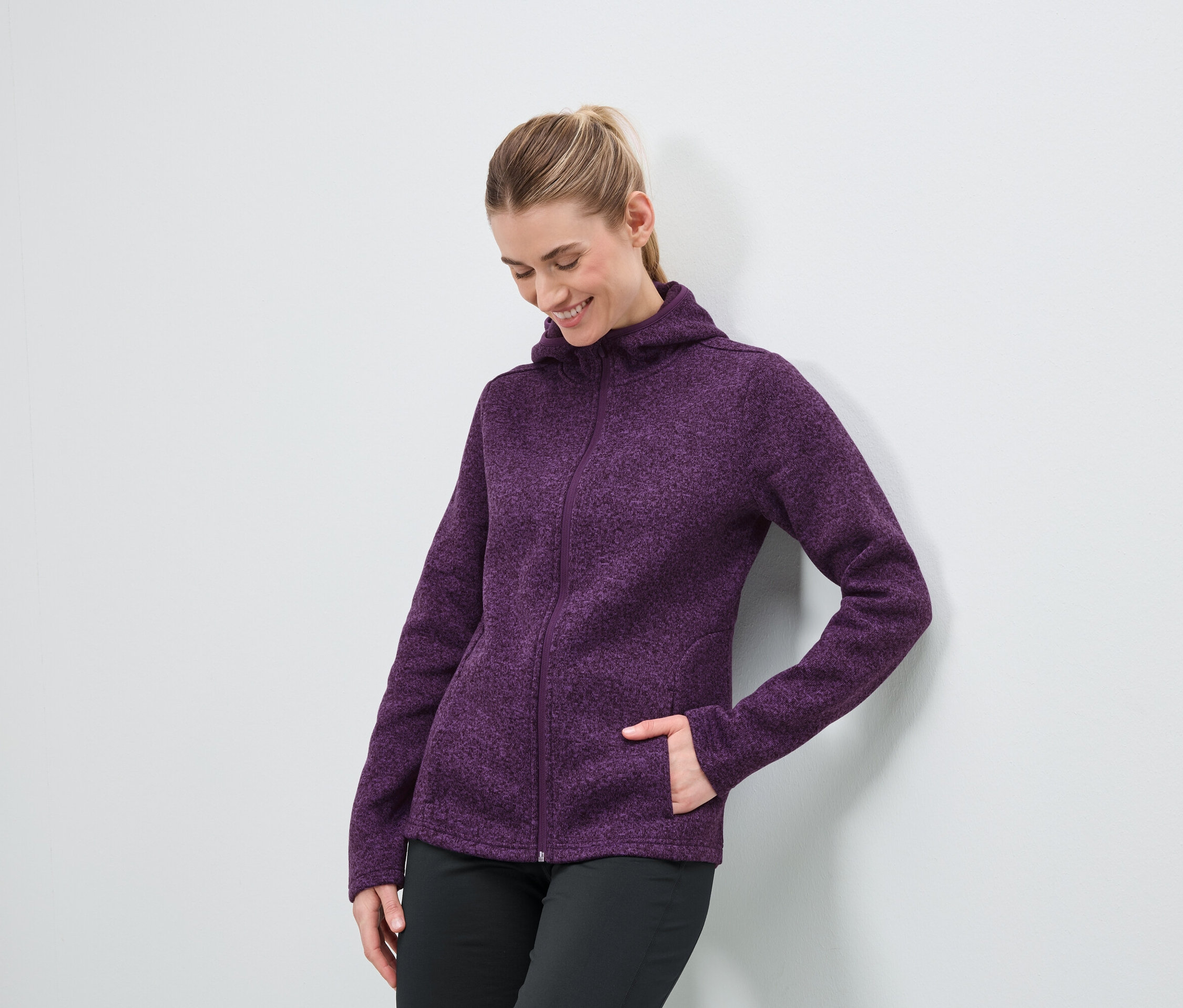 Une femme portant un sweat à capuche zippé violet et un pantalon noir se tient contre un mur blanc.