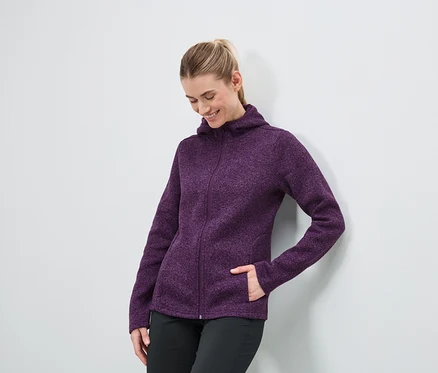 Une femme portant un sweat à capuche zippé violet et un pantalon noir se tient contre un mur blanc.