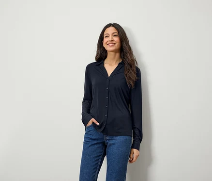 Une femme souriante aux longs cheveux bruns pose en chemise bleue à manches longues et en jean.
