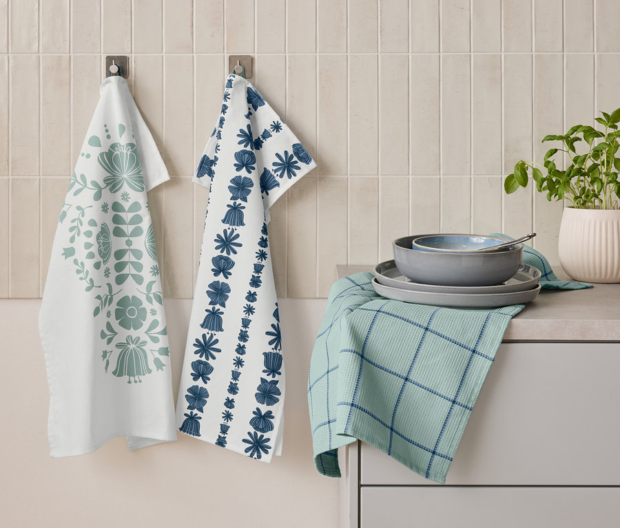 Deux torchons à motifs floraux sont accrochés côte à côte au mur. En dessous, un torchon vert plié à rayures bleues, sur lequel sont empilées des assiettes et un bol. À côté se trouve un pot avec des herbes.