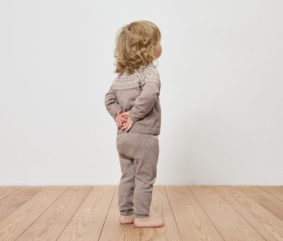 Un enfant vêtu de vêtements en tricot marron se tient dos à la caméra sur un plancher en bois.