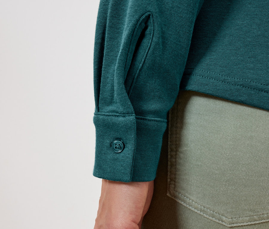 Détail d'une chemise verte à manches longues avec un bouton sur la manchette et d'un jean vert olive.