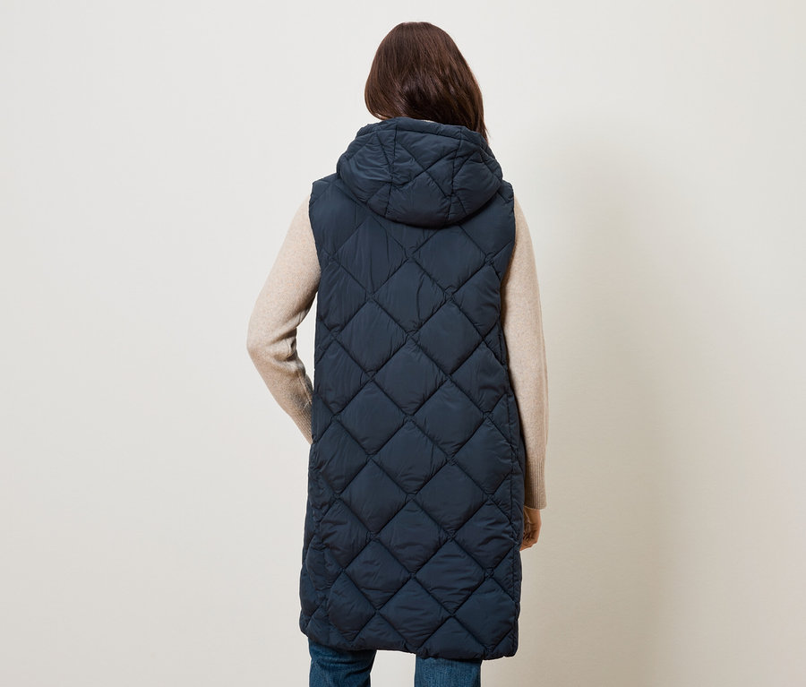 Une femme, vue de dos, porte une longue veste sans manches bleu marine matelassée avec une capuche.