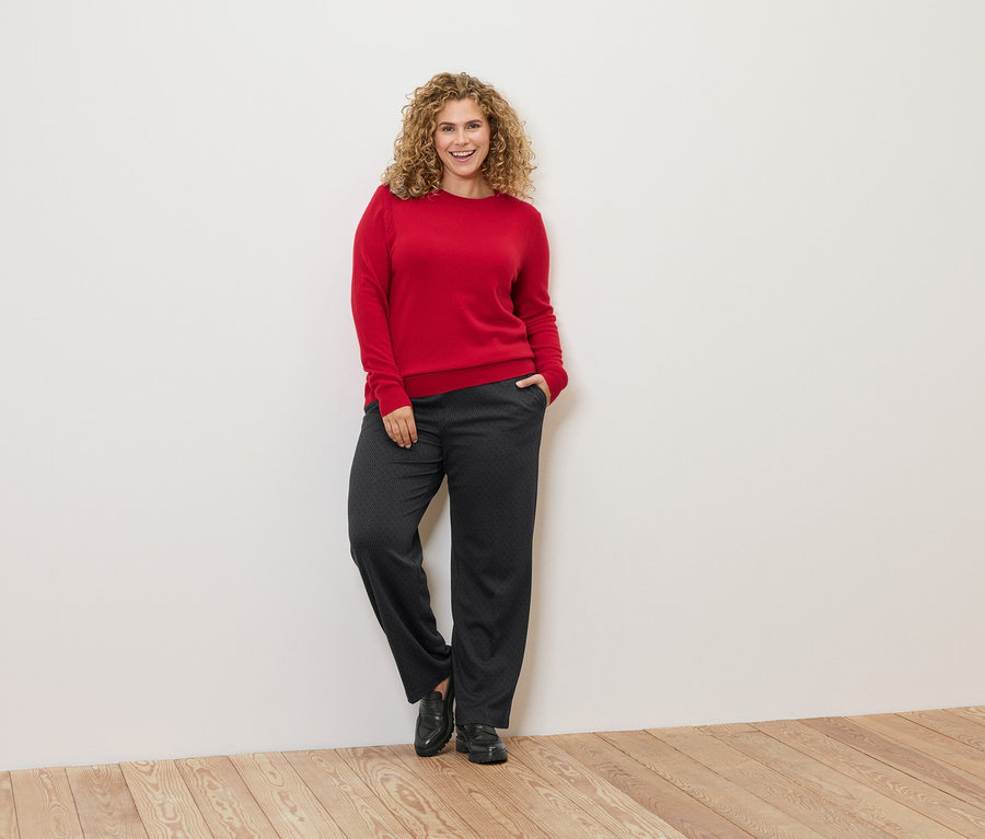 Une femme aux cheveux bouclés est appuyée contre un mur et porte un pull rouge et un pantalon foncé.