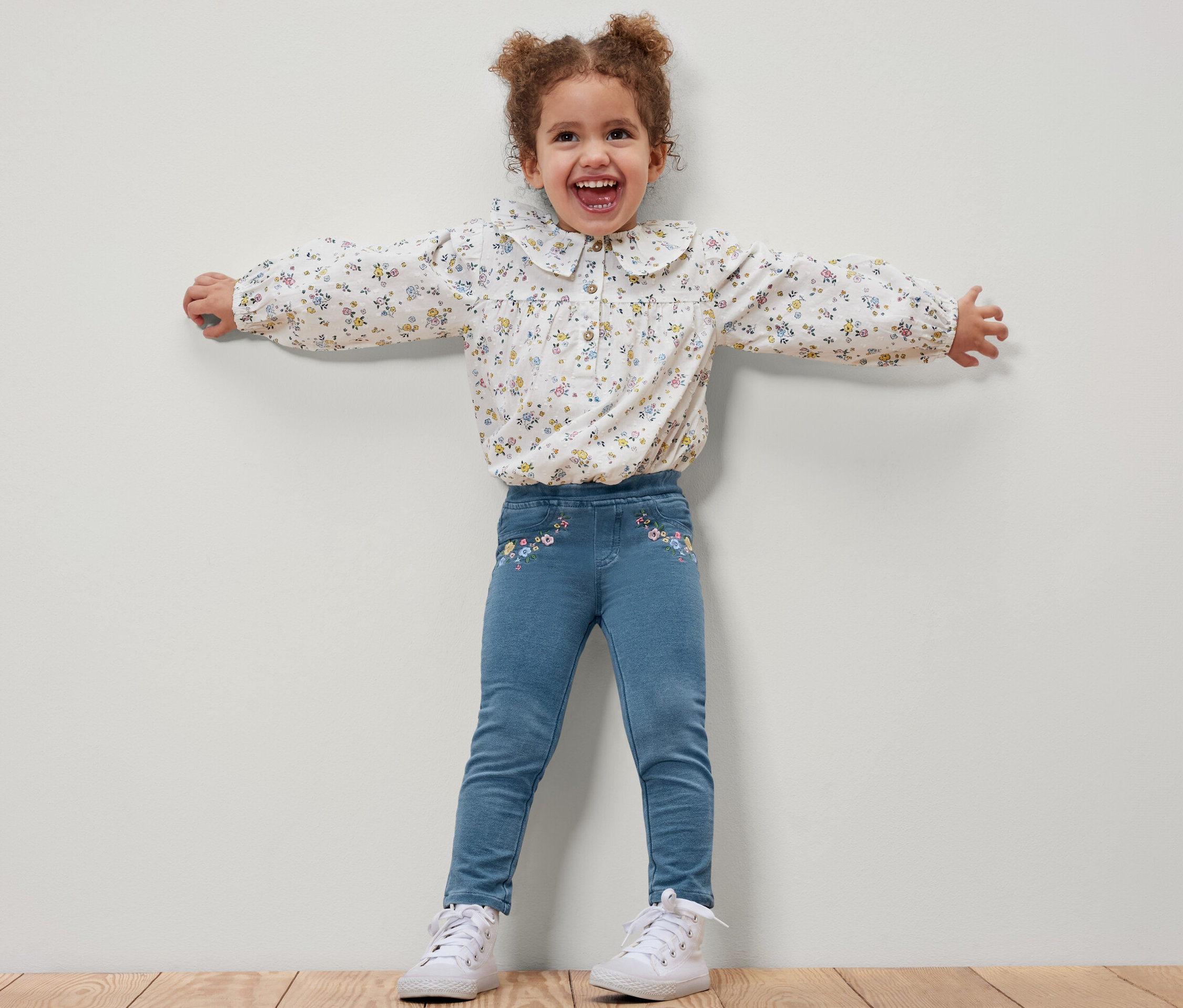 Une petite fille souriante se tient debout, les bras écartés, et porte un jegging pour enfant – Fit « Mia » et un chemisier à fleurs.
