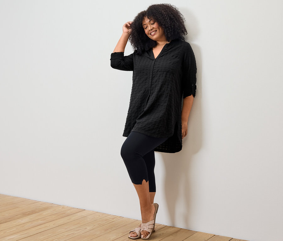 Femme posant en tunique longue noire à structure tissée et legging 3/4 avec détail.