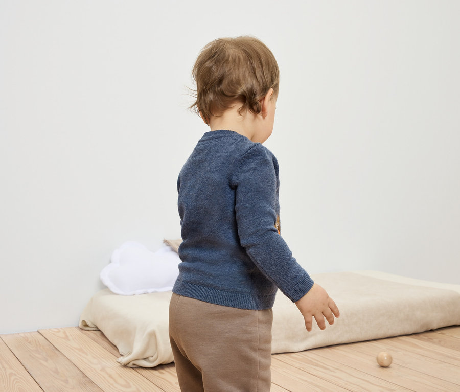 Un bébé vêtu d'un pull bleu et d'un pantalon marron se tient dos à l'objectif et joue avec un jouet en bois.