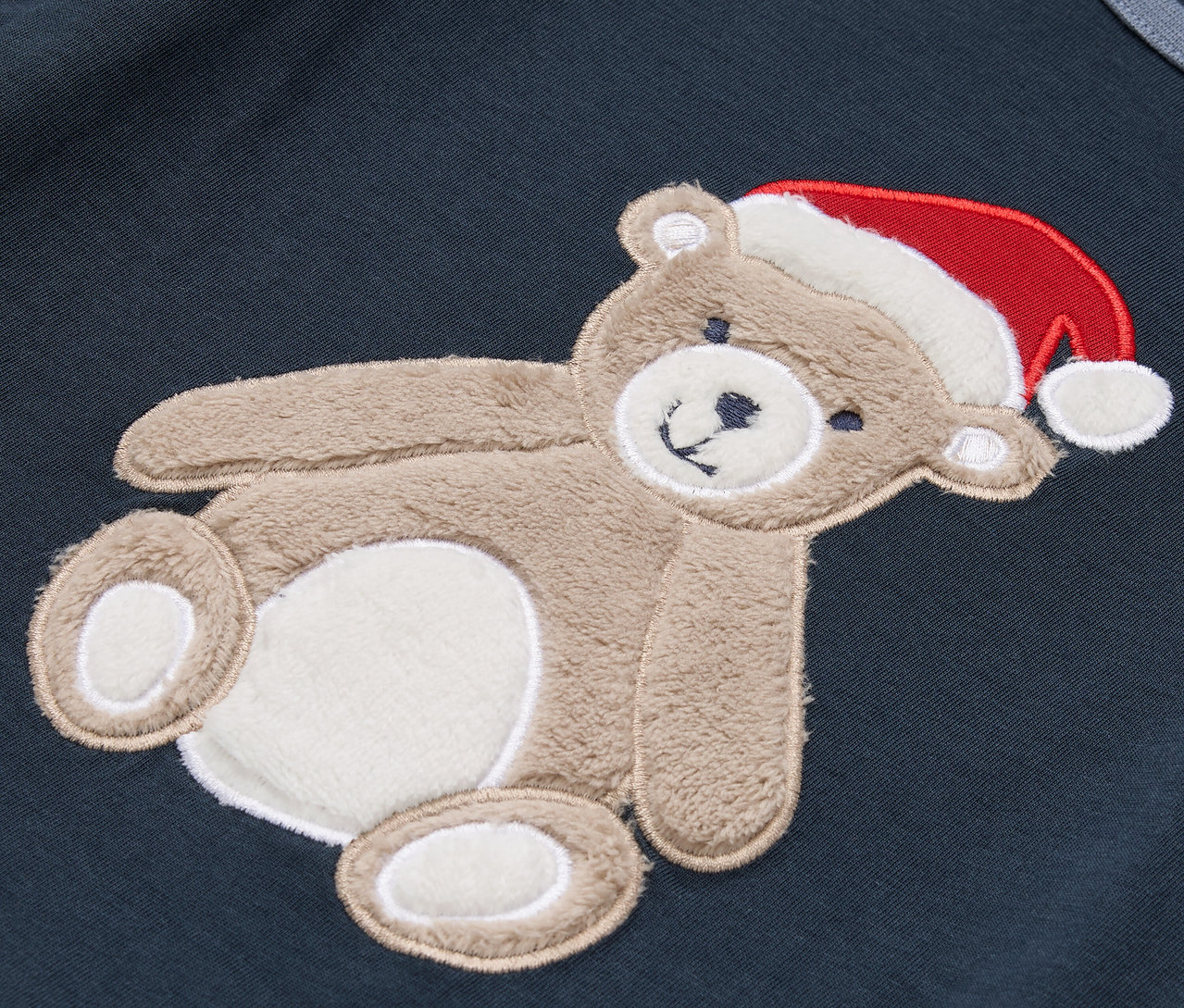 Sur un tissu bleu foncé se trouve un ours en peluche marron avec un bonnet de Noël rouge.