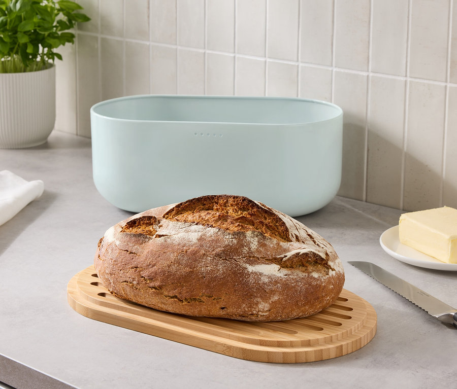 Un pain sur une planche à découper en bois à côté d'une boîte à pain bleue, une portion de beurre et un couteau sur un comptoir.