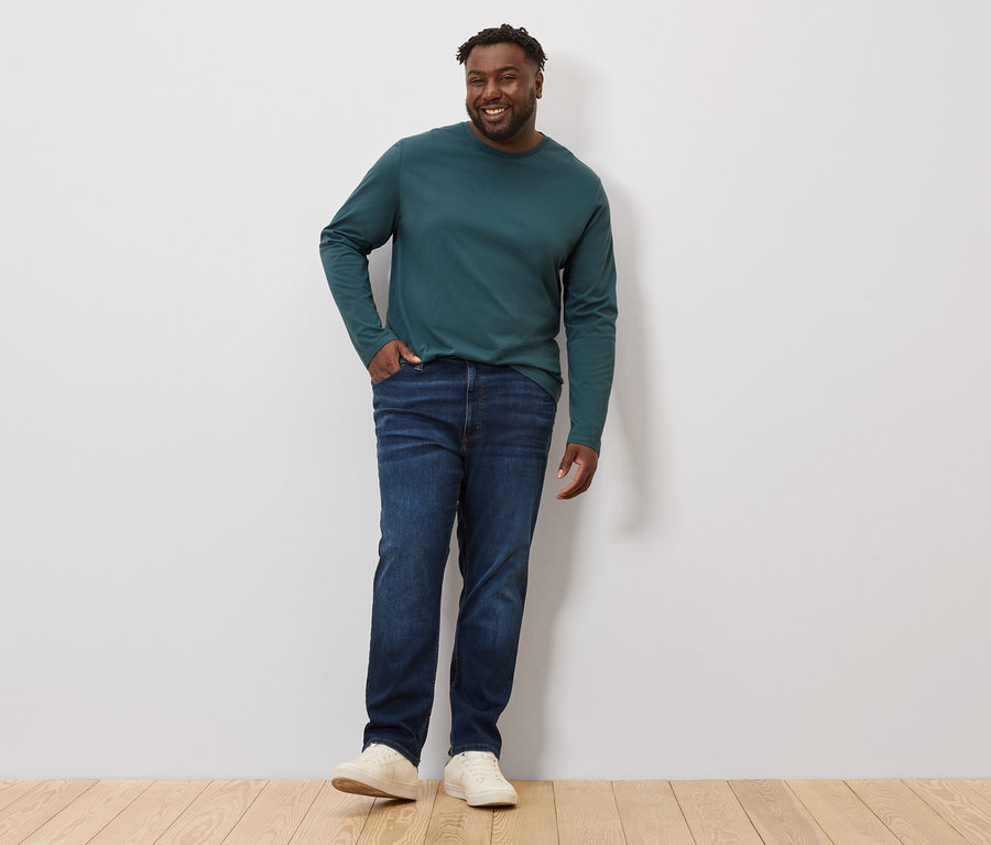 Un homme pose, vêtu d'un t-shirt vert à manches longues et d'un jean. Il porte 2 t-shirts à manches longues.