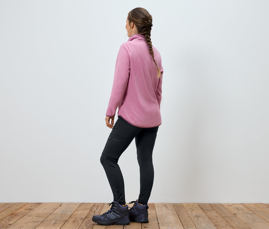 Une femme portant une veste en polaire rose, un pantalon noir et des bottes, se tient dos à la caméra.