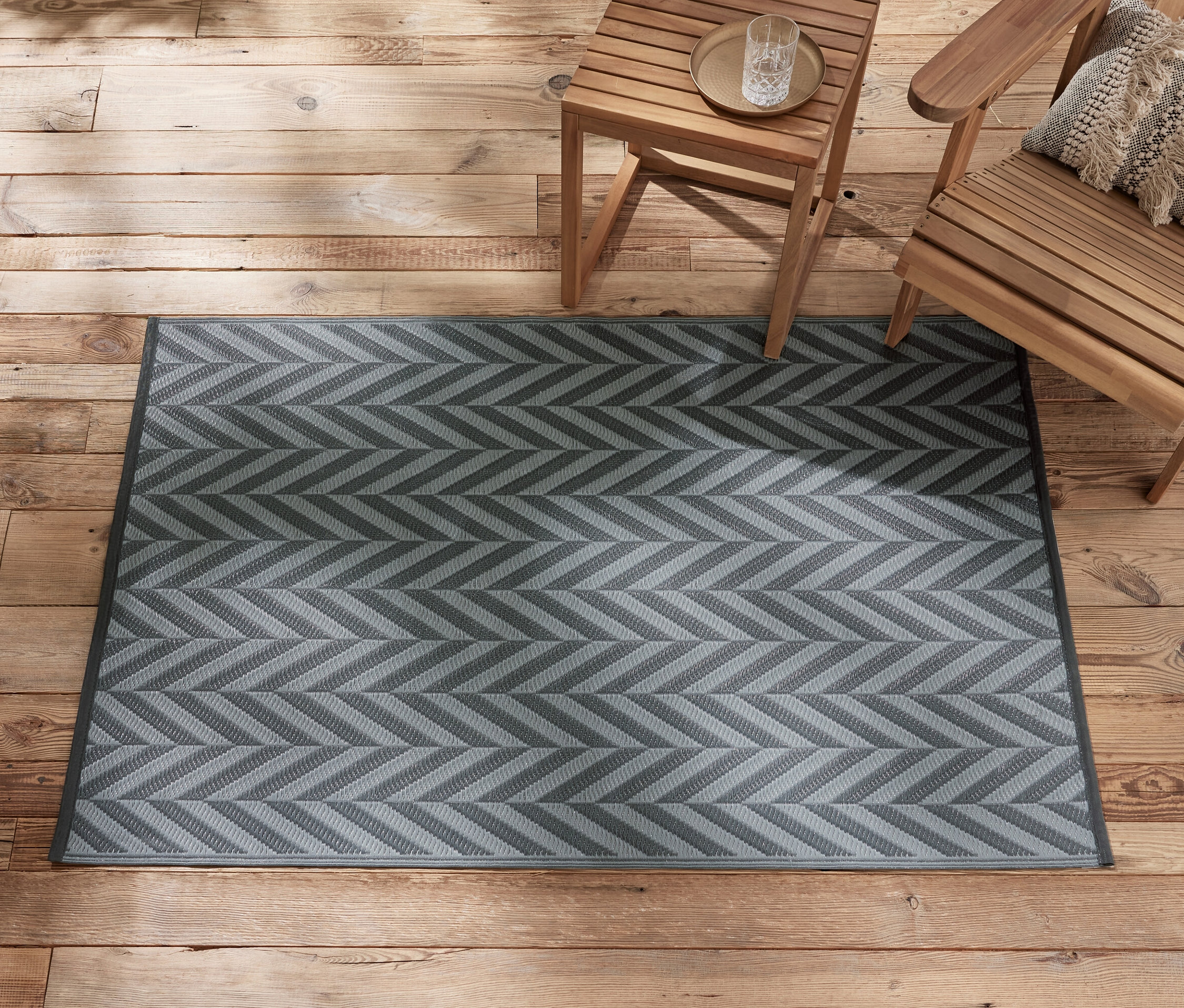Un tapis d’extérieur anthracite-gris clair d’environ 165 x 115 cm est posé sur un plancher en bois à côté de meubles de jardin en bois.