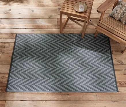 Un tapis d’extérieur anthracite-gris clair d’environ 165 x 115 cm est posé sur un plancher en bois à côté de meubles de jardin en bois.