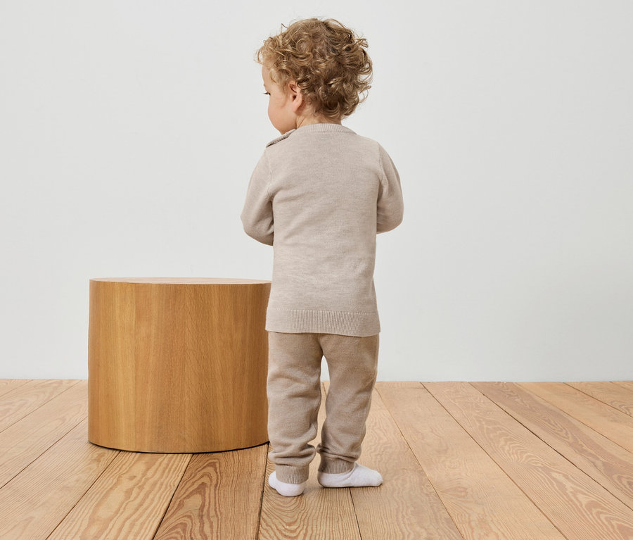 Un bébé est vu de dos, portant un pull et un pantalon beiges, à côté d'une petite table en bois.