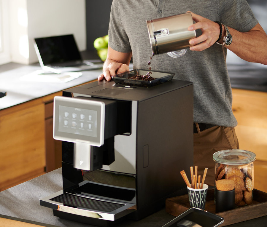 Homme avec des lunettes versant des grains de café dans une cafetière automatique Tchibo Office noire.