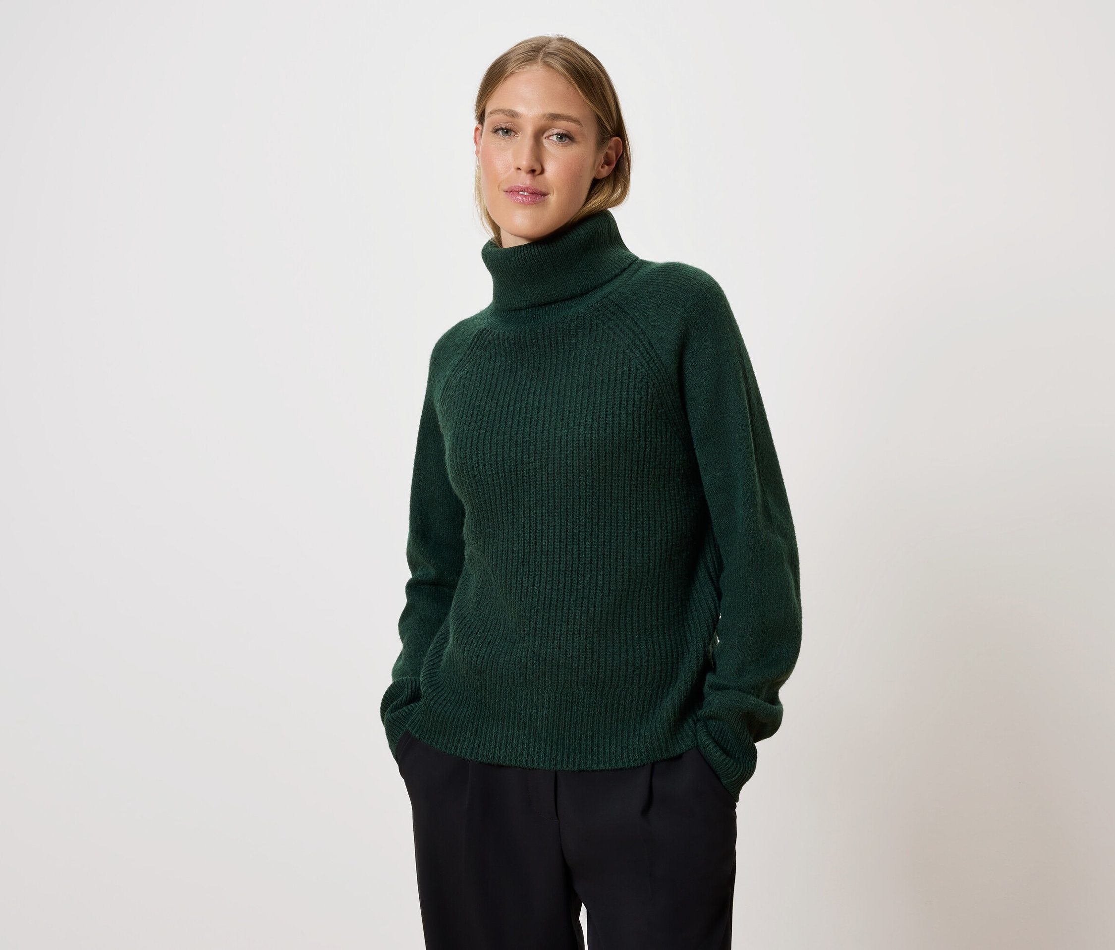 Femme posant dans un pull à col roulé vert et un pantalon noir.