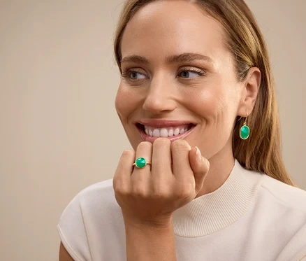 Femme souriante portant une bague avec une pierre verte à l'annulaire et des boucles d'oreilles pendantes avec une pierre verte.