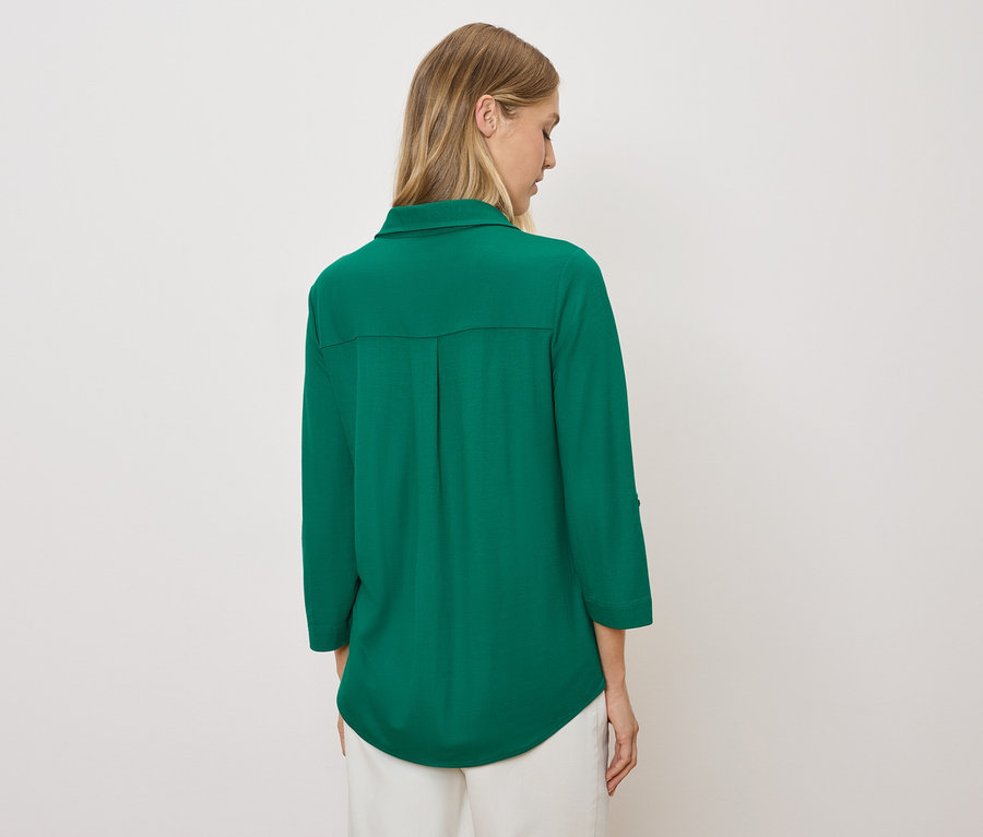 Une femme, de dos, porte une chemise verte et un pantalon blanc.