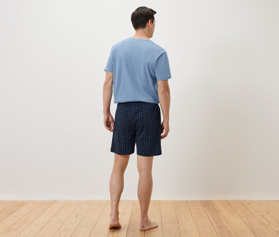 Un homme se tient dos à la caméra, vêtu d'un t-shirt bleu et d'un short bleu marine à motifs.