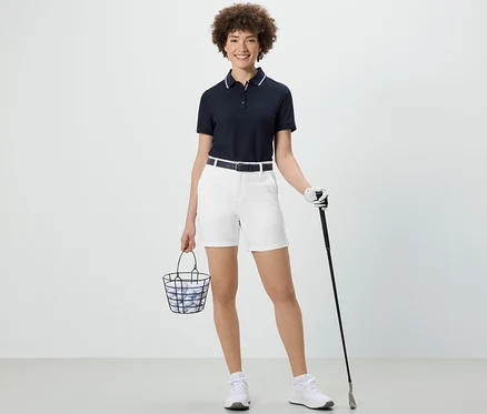 Femme tenant un panier de balles de golf et un club de golf. Elle porte un short technique en softshell blanc.